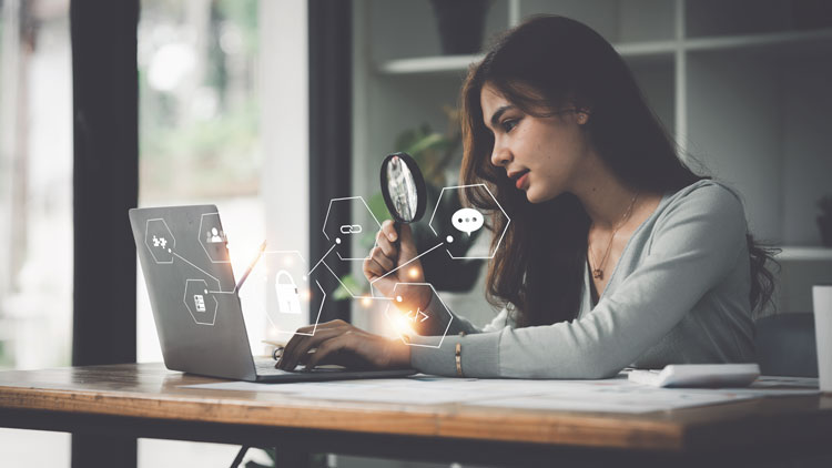 Frau sitzt vor einem Laptop und blickt durch eine Lupe auf den Bildschirm aus dem Sicehrheitssymbole erscheinen; Copyright Panthermedia